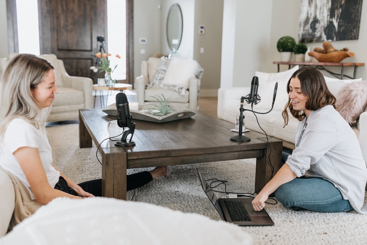 Two ladies record audio together on a laptop with a microphone each