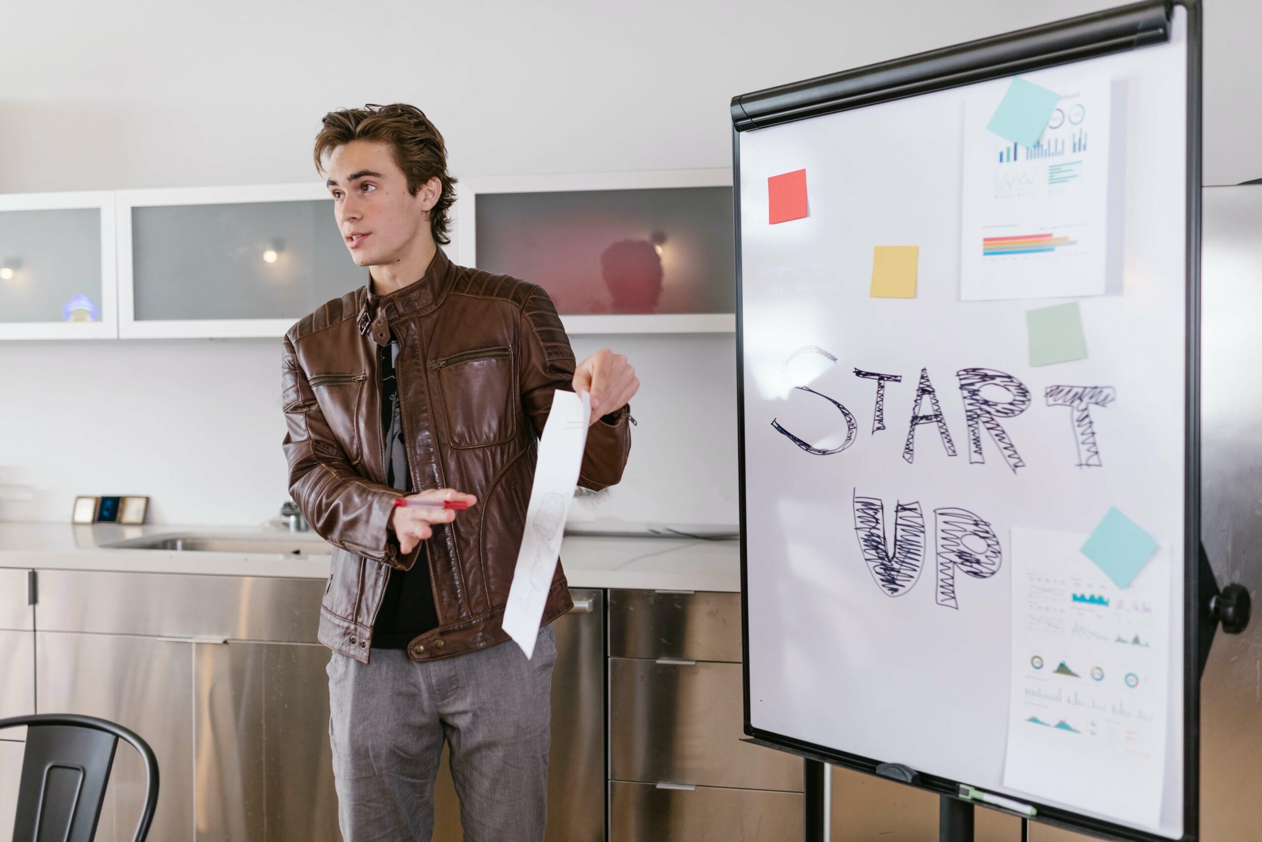 A man in a brown leather jacket gives a presentation standing next to a whiteboard which says "start up"