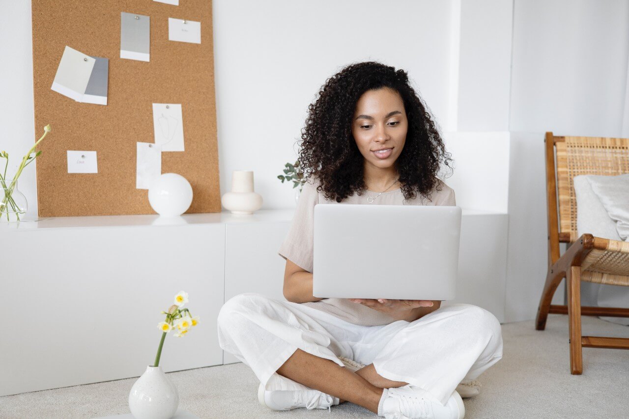 Woman with afro hair wearing white trousers sits cross legged on floor working on laptop