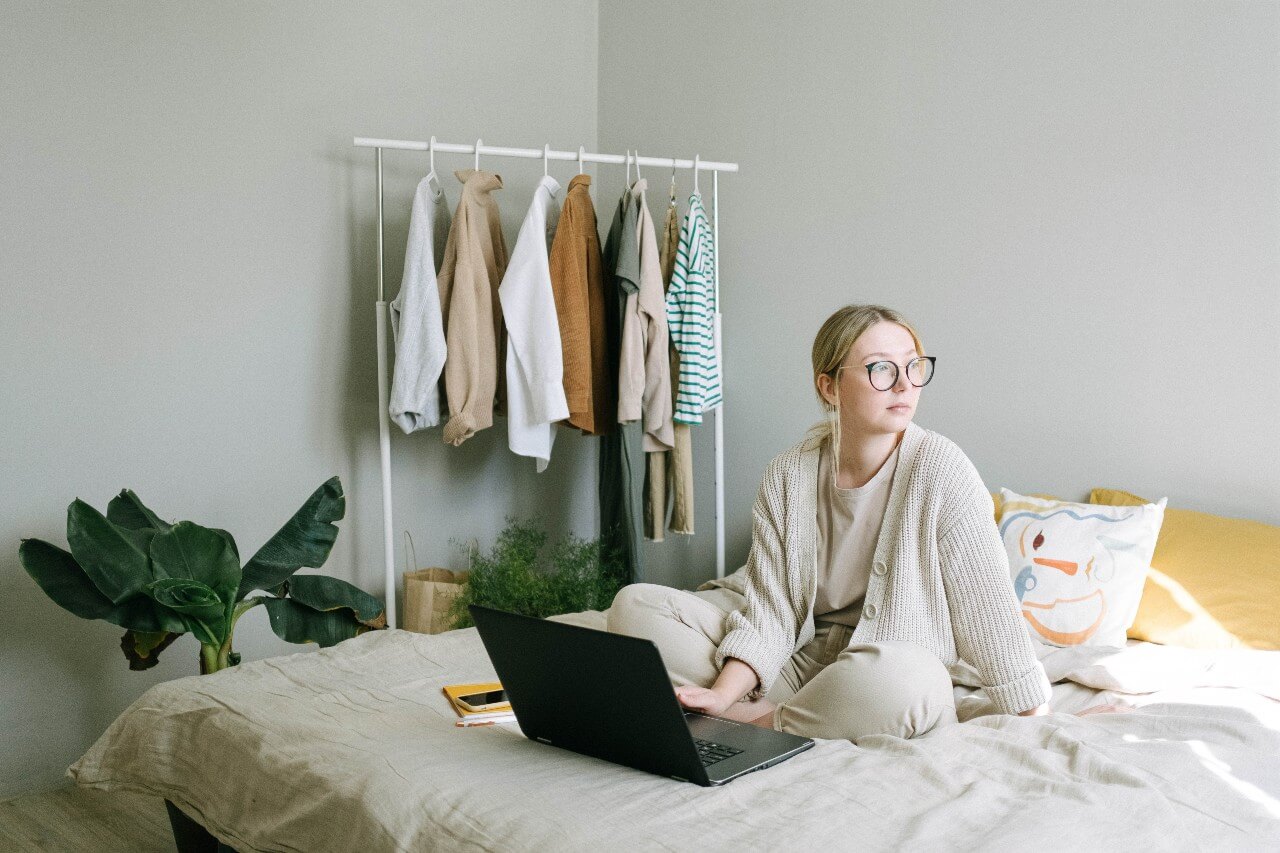 Woman in cream cardigan sits on bed working on laptop