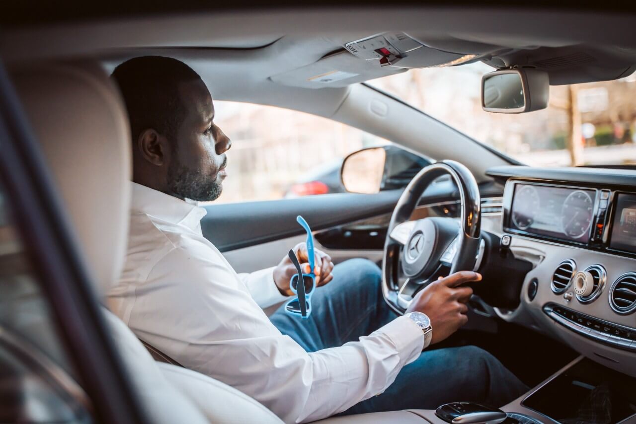 Featured image for HMRC mileage rates post. Image shows black man wearing white shirt and blue jeans sitting in the drivers seat of a Mercedes car.