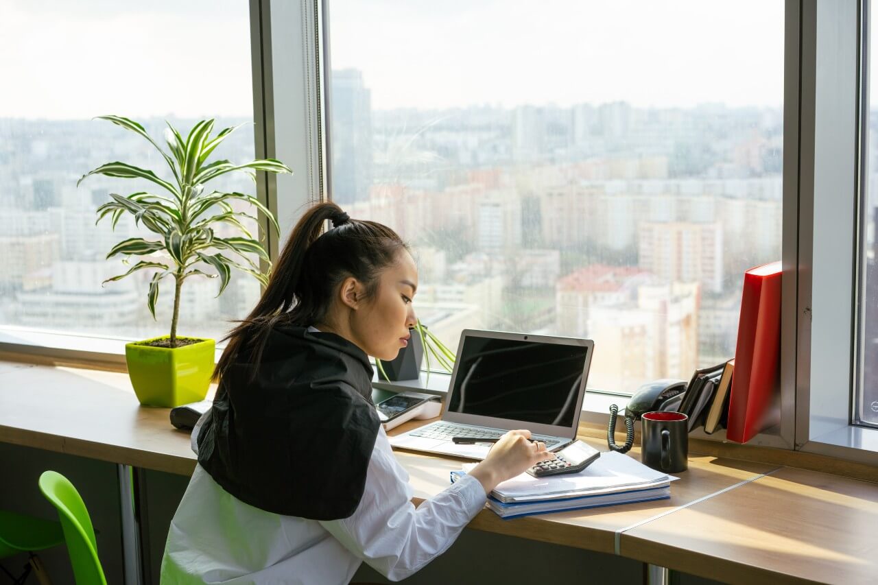 A woman sits at a window desk working at a laptop, working out calculations with a calculator