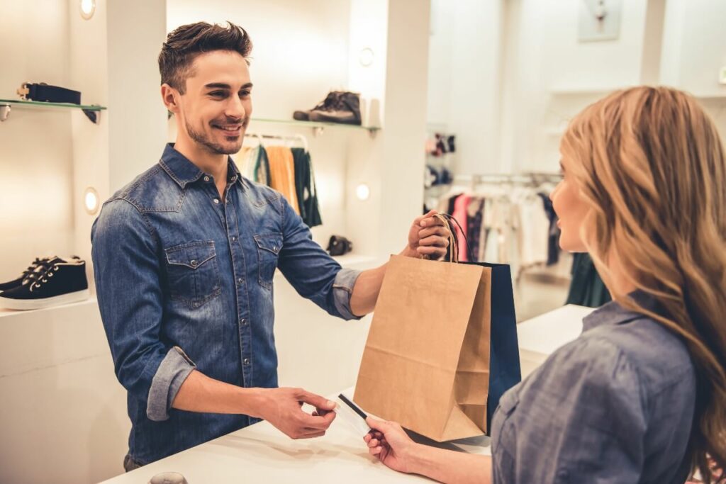 A man wearing a denim shirt hands a lady a bag at the payment counter of a retail store