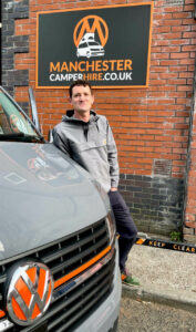 Nick, owner of Manchester Camper Hire stands in front of sign that reads manchestercamperhire.co.uk