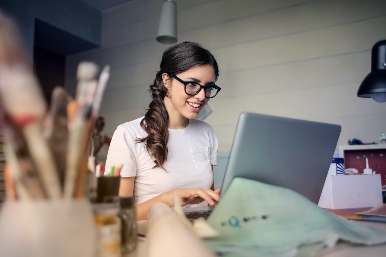 Woman with long brown hair in a ponytail wearing glasses sits at a desk working on a laptop. There is a pot of paintbrushes beside her.