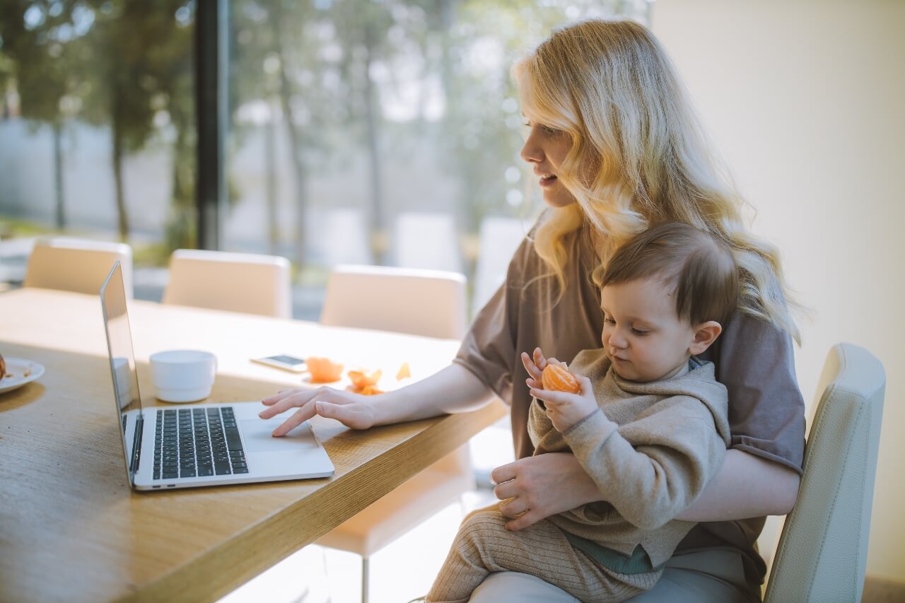 A lady works at her laptop with a young child on her lap