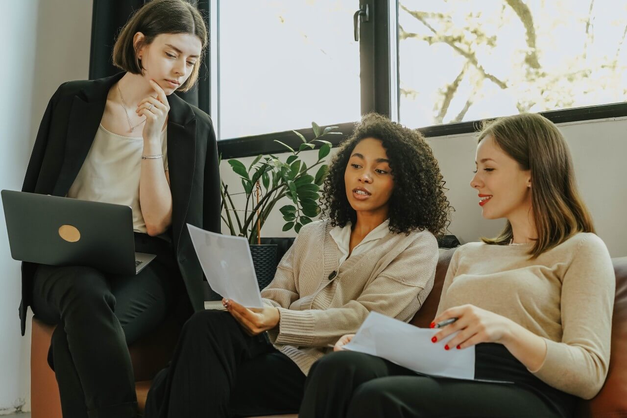 Three women sit working together side by side on a sofa