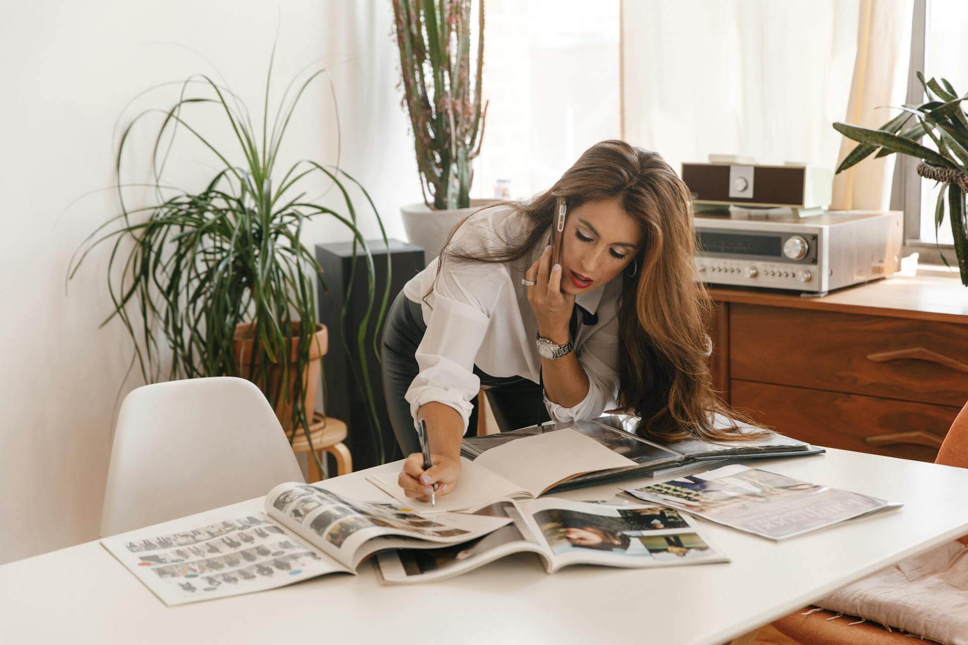 a female small business owner with brown hair wearing a white shirt holding a phone to one ear while she works on her fashion magazine.