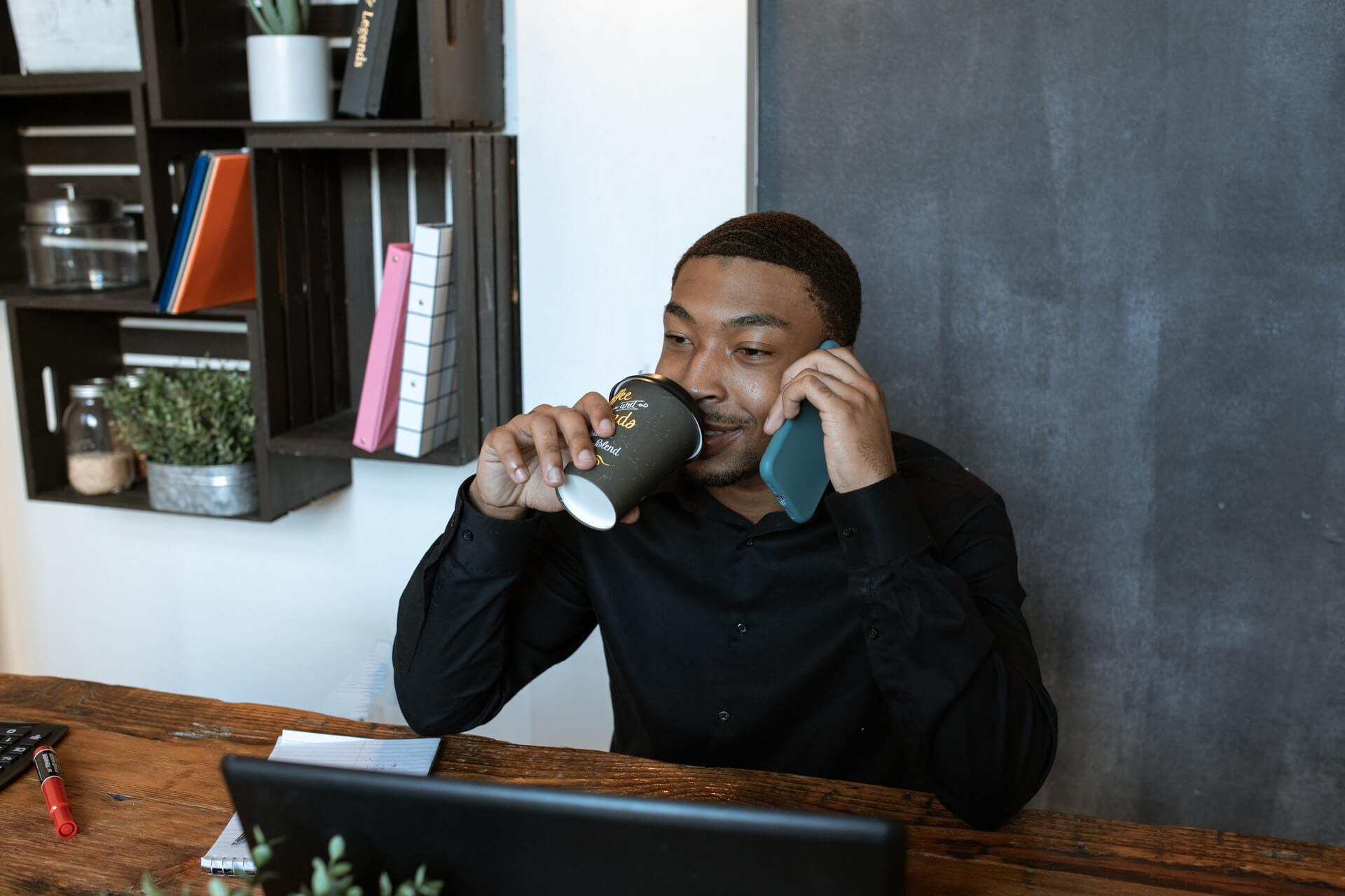 A man looks at his laptop screen while drinking a coffee and speaking on the phone