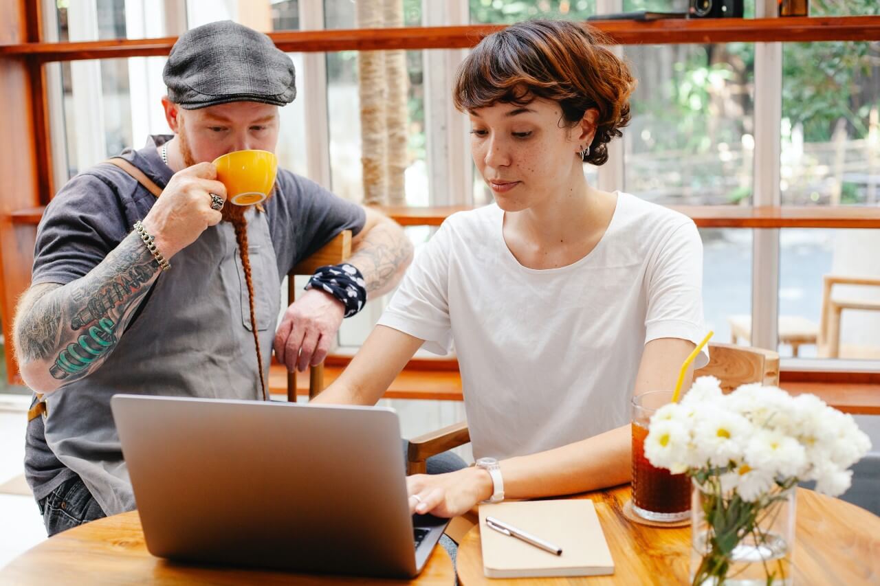 A woman in a white top shows a man wearing a cap and drinking a cup of tea something on her laptop screen