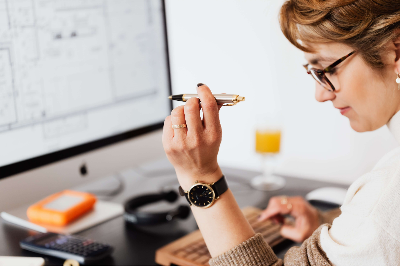 A lady holding a pen works at her computer doing calculations with a calculator